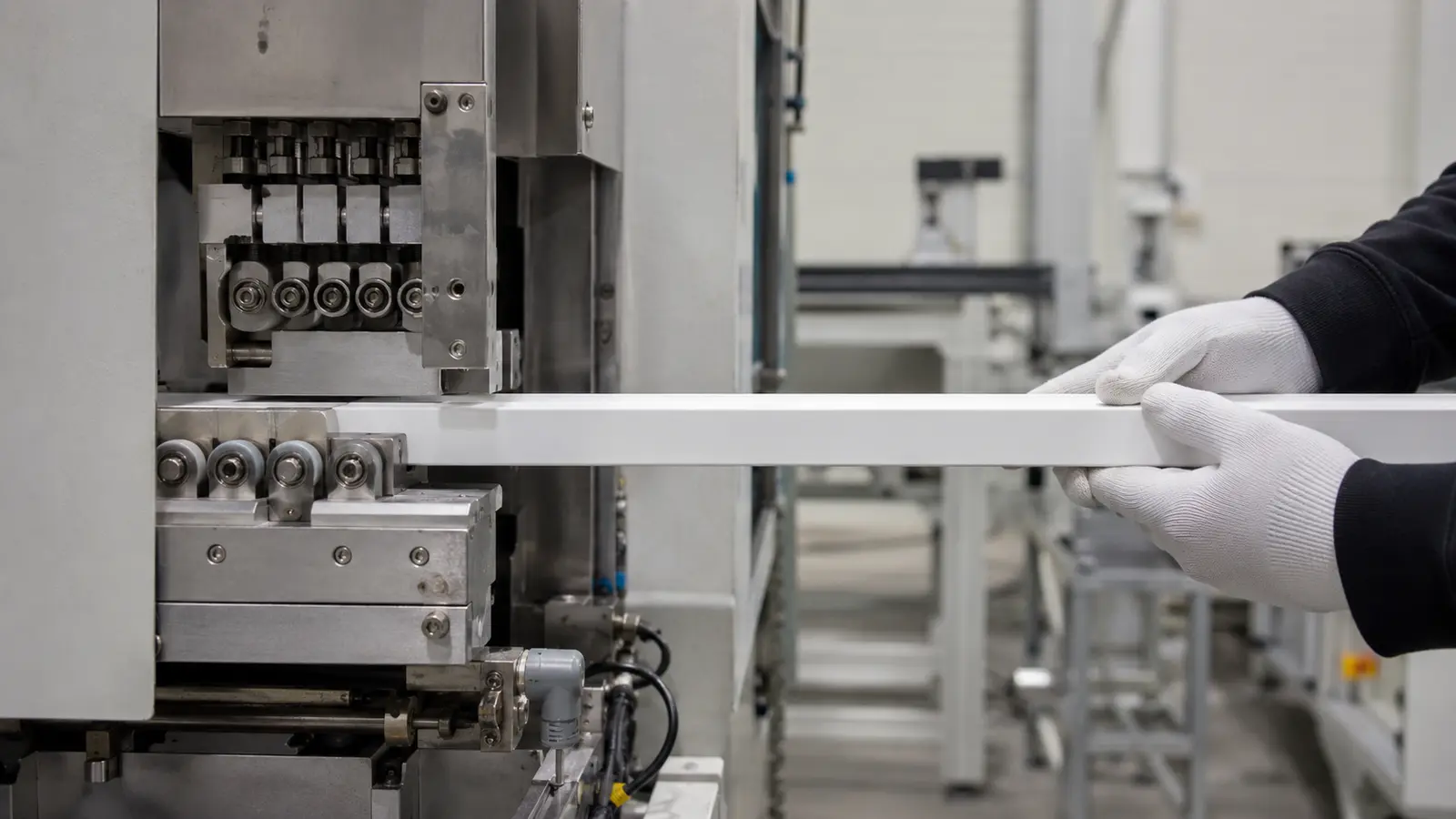 Gloved hands guiding a white lighting profile through production machinery.